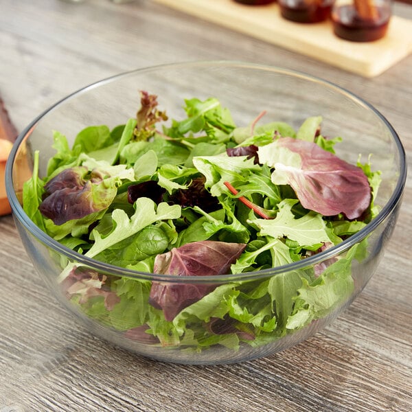 A clear glass bowl filled with leafy green salad, placed on a wooden table.