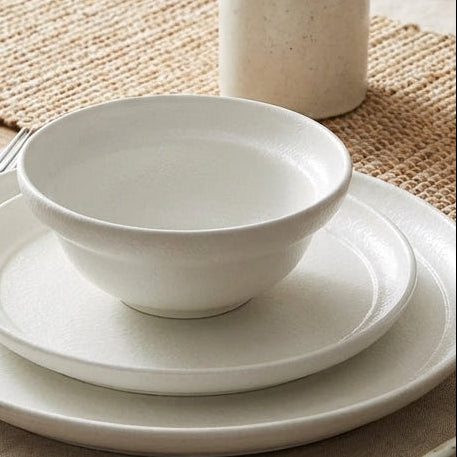 White ceramic tableware set on a wooden table with a vase and placemats.