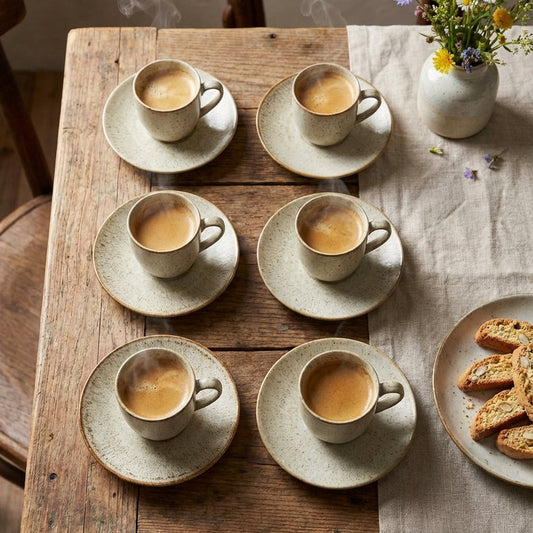Set of six ceramic coffee cups and saucers on a wooden table with a vase of flowers and a plate of cookies.