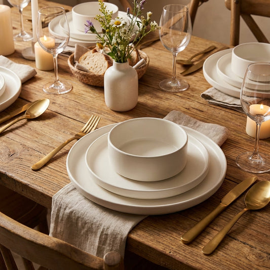 Dining table setting with white plates, gold cutlery, and candles on a wooden table.
