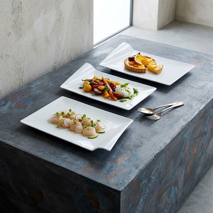 Three white rectangular ceramic plates with wavy edges are arranged on a rustic blue-gray stone table, styled with plated food and silver cutlery in a bright interior setting.
