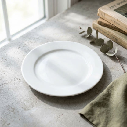 A white ceramic plate with a wide rim, styled on a textured gray stone surface next to a green linen napkin, eucalyptus stems, and vintage books.