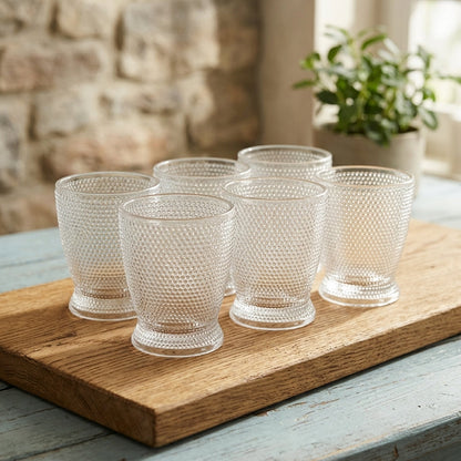 Five clear textured glass tumblers arranged on a wooden serving board on a rustic table, with a stone wall and potted plant in the background.