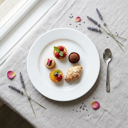 A white ceramic plate styled on a linen tablecloth with lavender sprigs, rose petals, and a silver spoon, holding four assorted pastries including tartlets, a chocolate truffle, and a croissant.