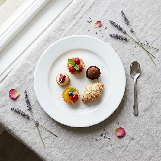 A white ceramic plate styled on a linen tablecloth with lavender sprigs, rose petals, and a silver spoon, holding four assorted pastries including tartlets, a chocolate truffle, and a croissant.