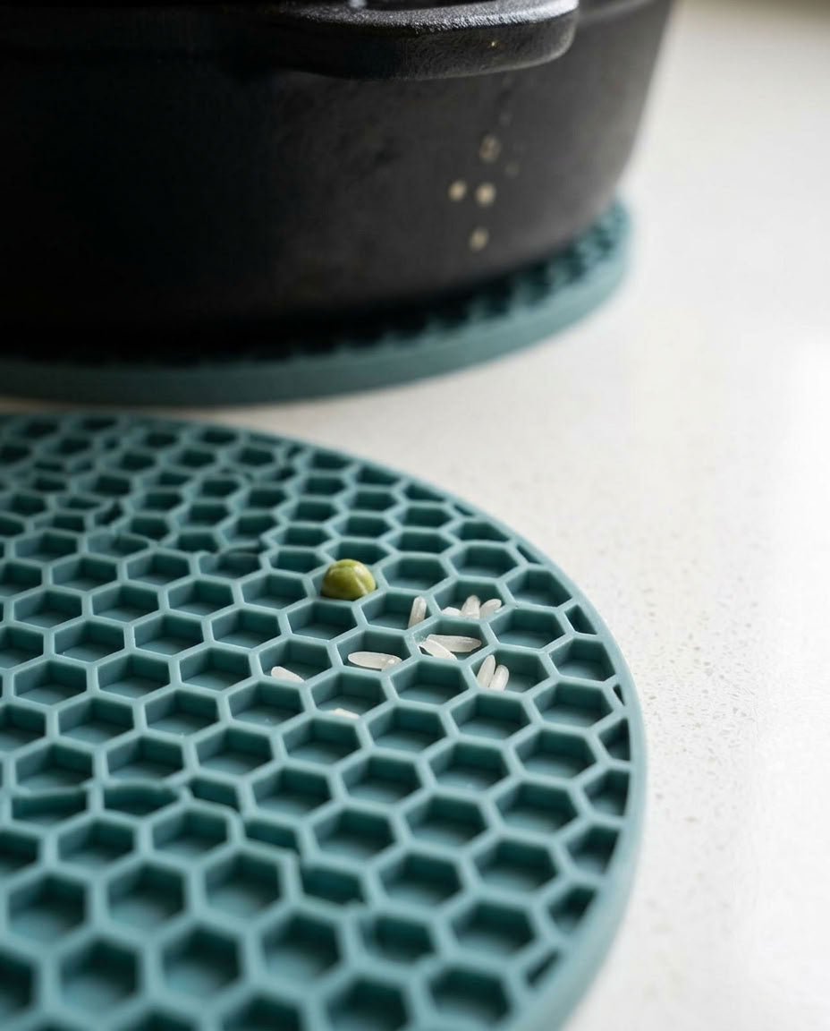 A close-up of a teal silicone trivet with a honeycomb pattern, shown on a light countertop with a pot partially visible in the background.