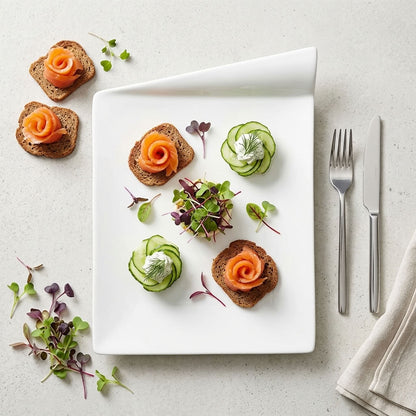 A white rectangular serving platter with a folded corner, styled with four small appetizers: two topped with orange salmon rosettes and two with cucumber rosettes, on a light stone surface. A fork and knife are placed to the right, with microgreens scattered around the platter.