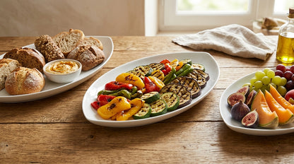 Plates of grilled vegetables, bread, and fruit on a wooden table.