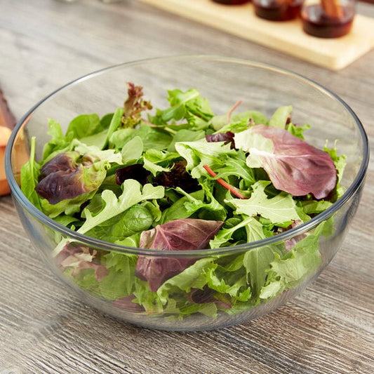 A clear glass bowl filled with leafy green salad, placed on a wooden table.