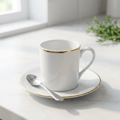 White mug with gold rim on a saucer with a spoon, placed on a light surface with a window and plant in the background.