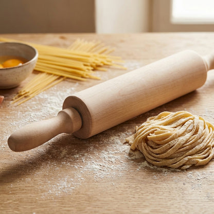 A light natural wood rolling pin resting on a wooden table dusted with flour, next to a small pile of handmade pasta and a small bowl with an egg yolk. Uncooked spaghetti is visible in the background.