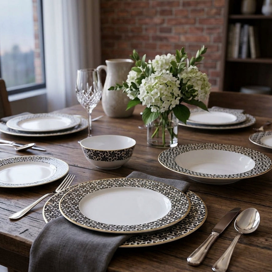 Dining table set with patterned plates, silverware, and a vase of flowers in a room with large windows.
