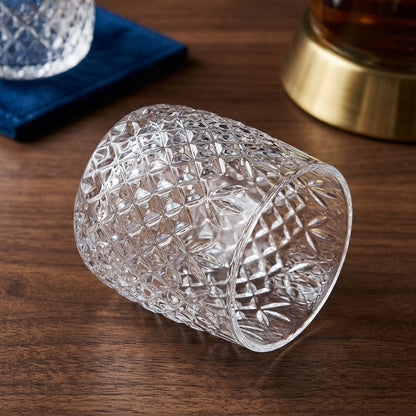 A clear cut-crystal tumbler glass with a diamond pattern, lying on its side on a dark wooden table.