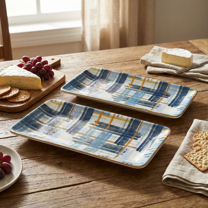 Two plaid ceramic trays on a wooden table with grapes, cheese, and crackers.