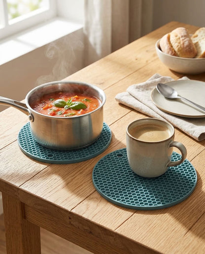 Two round teal silicone trivet coasters on a wooden dining table, one supporting a pot of tomato soup and the other a ceramic mug of coffee. A plate, spoon, and bread bowl are visible in the background near a window.