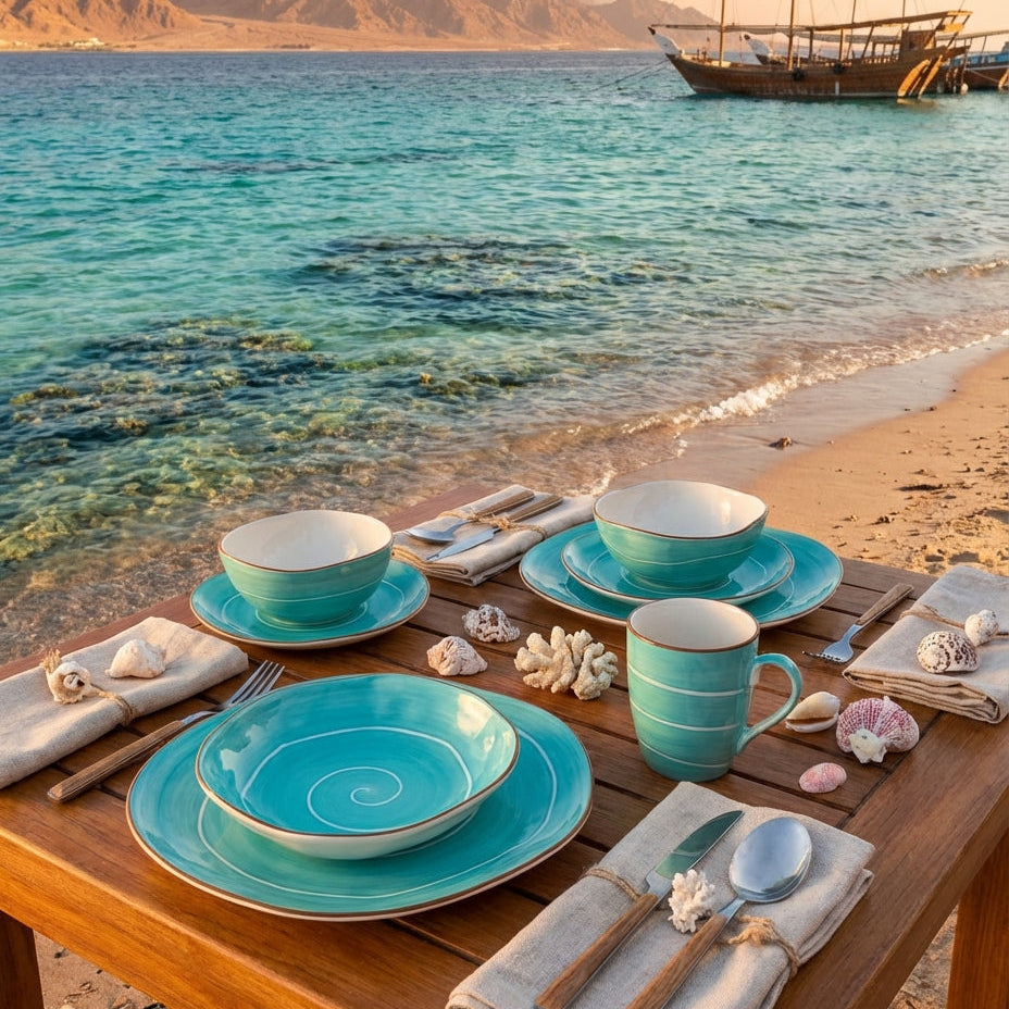 Table setting with turquoise dishes and cups on a beach with mountains and a boat in the background.