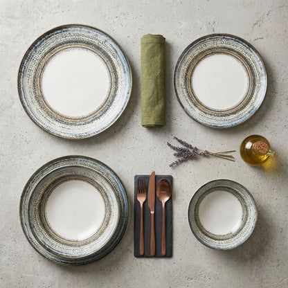 A styled flat-lay of a dinnerware set on a light stone surface, featuring four plates of varying sizes with a rustic textured rim, a rolled olive green napkin, a small bottle of oil, and cutlery on a dark slate card.