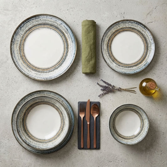 A styled flat-lay of a dinnerware set on a light stone surface, featuring four plates of varying sizes with a rustic textured rim, a rolled olive green napkin, a small bottle of oil, and cutlery on a dark slate card.