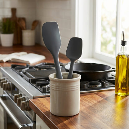 Gray silicone spatulas in a white ceramic holder on a kitchen counter with a stove and window in the background.