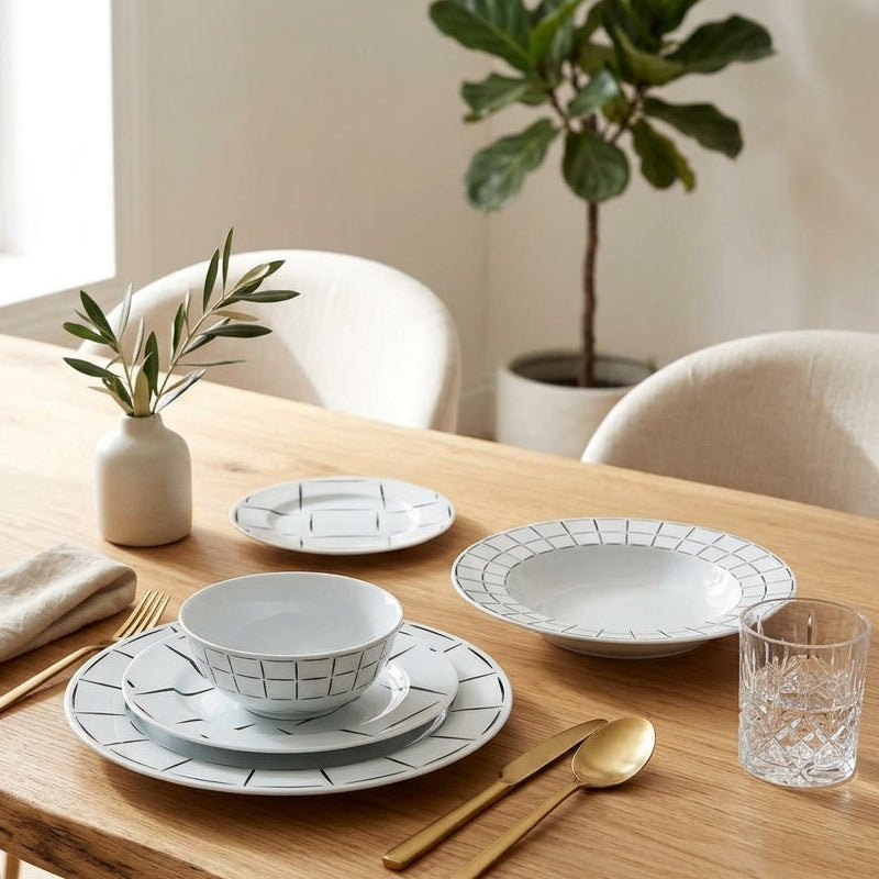 Dining table set with white ceramic plates, gold cutlery, and a plant in a bright room.