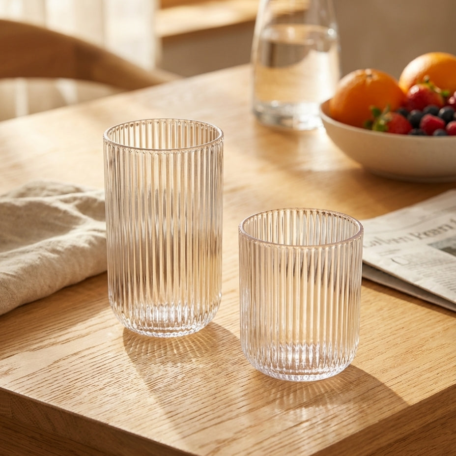 Two clear glass tumblers on a wooden table with a bowl of fruit and newspaper in the background.
