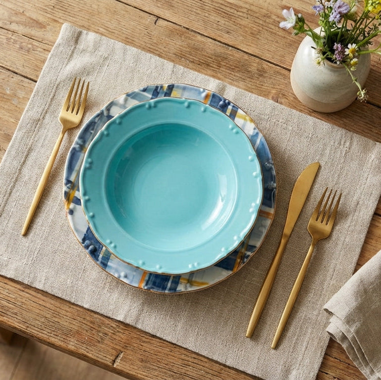 Teal plate with decorative rim on a wooden table with gold cutlery and a vase of flowers.