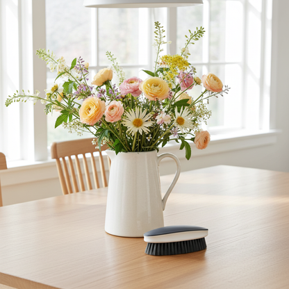 White pitcher with a bouquet of flowers on a wooden table, with a brush next to it.
