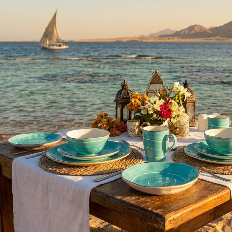 Dining table set with turquoise dishes and cups on a beach with ocean and sailboat in the background
