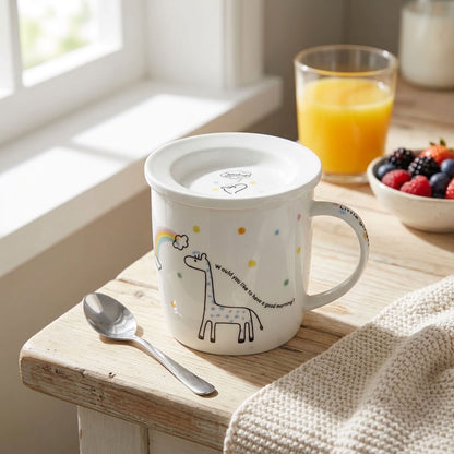 A white ceramic mug with a lid, featuring a cute giraffe illustration, a small rainbow, colorful dots, and the text 'we could all like a hot good morning!' on a wooden table by a window. A silver spoon rests beside the mug, with a glass of orange juice and a bowl of fresh berries in the background.