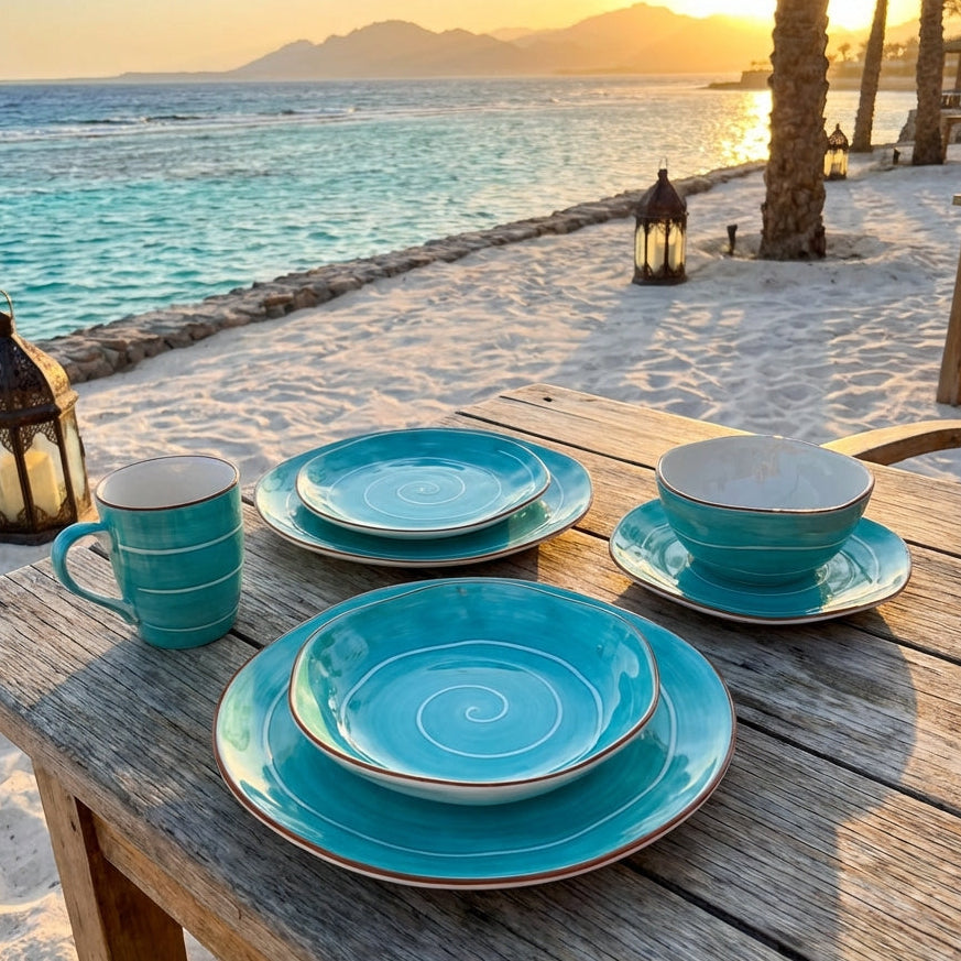 Turquoise ceramic tableware set on a wooden table by the beach at sunset.