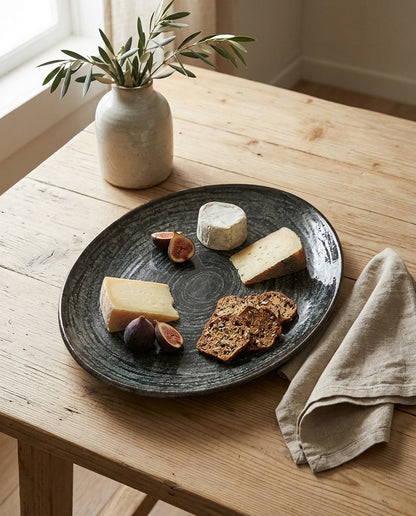 A round dark charcoal gray ceramic serving platter with a textured, hand-painted finish, styled with cheese, figs, and crackers on a light wood table next to a linen napkin and a ceramic vase.