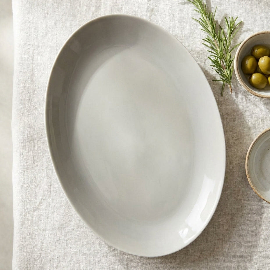 White oval ceramic plate on a light fabric background with a small bowl of olives and rosemary.