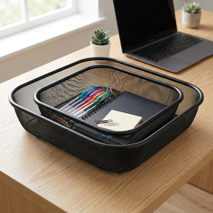 A black mesh desk organizer tray with a double-tray stack, shown on a light wood desk with a laptop, notebook, and small potted plants in the background.