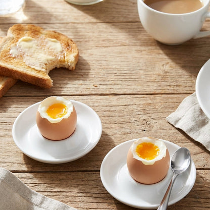 Two soft-boiled eggs on white plates with toast and coffee on a wooden table.
