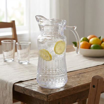 Clear glass pitcher with lemon slices on a wooden table with glasses and fruit in the background.