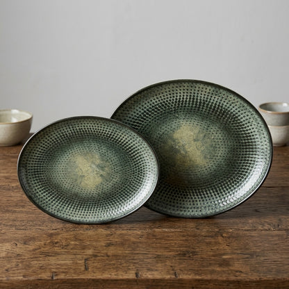 Two round ceramic serving platters with a textured, dimpled surface and a distressed olive green and beige glaze, displayed on a rustic wooden table.