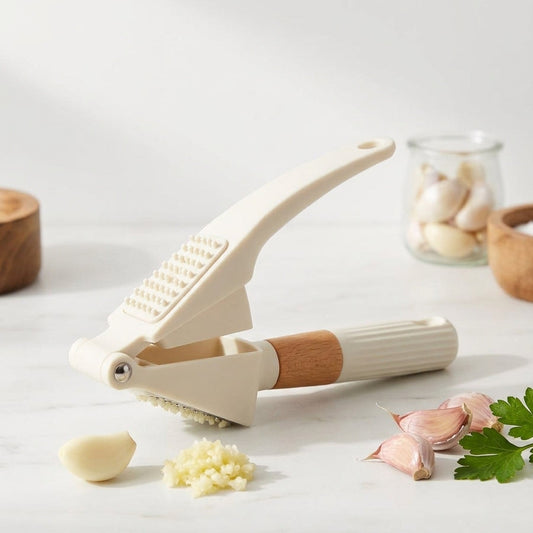 An off-white garlic press with a light brown wooden handle grip, shown on a white marble surface with garlic cloves, minced garlic, parsley, and wooden bowls in the background.