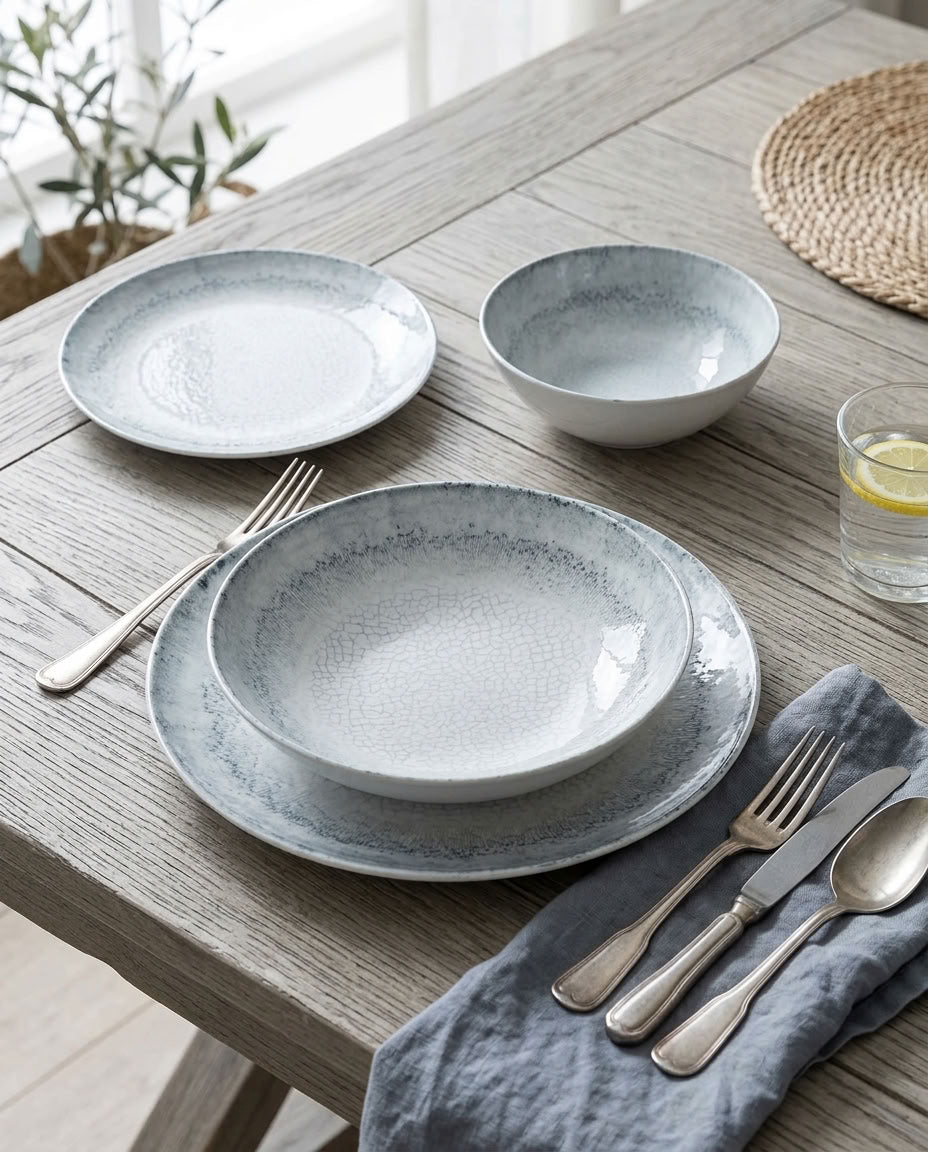 A place setting on a rustic wooden table featuring a textured light gray and white dinnerware set with matching silver cutlery and a blue-gray napkin.