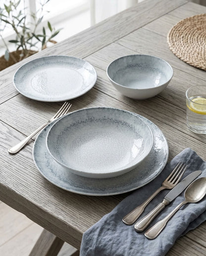 A place setting on a rustic wooden table featuring a textured light gray and white dinnerware set with matching silver cutlery and a blue-gray napkin.