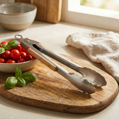 Metal tongs on a wooden cutting board with cherry tomatoes and basil leaves.