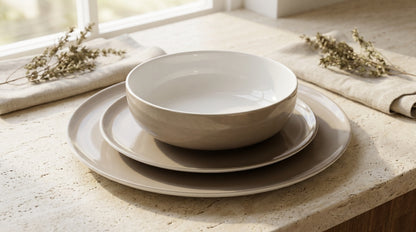 Set of beige ceramic dishes on a light wooden surface with a blurred window background.