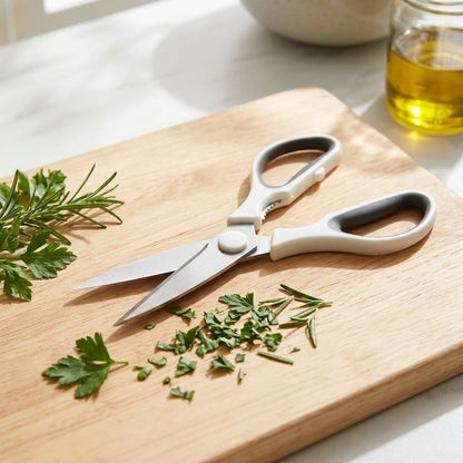 A pair of kitchen shears with white handles and silver blades resting on a light wood cutting board, surrounded by chopped parsley and rosemary, with a bowl of lemons and a glass of oil in the softly blurred background.