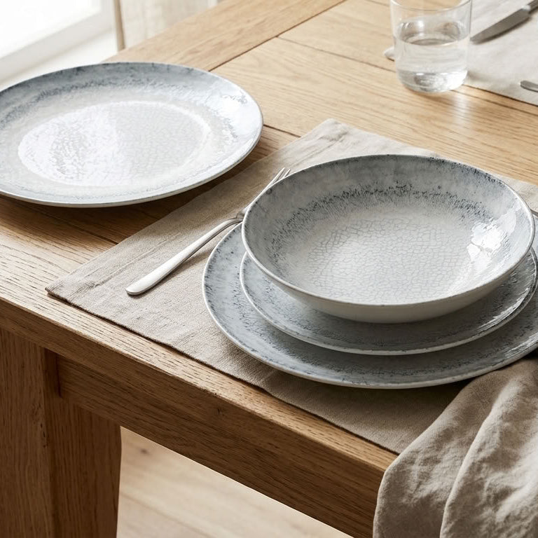 A rustic dining table setting featuring a coordinated ceramic tableware collection with textured speckled gray and off-white plates and bowls, styled with linen placemats, silver cutlery, and a small ceramic bottle.