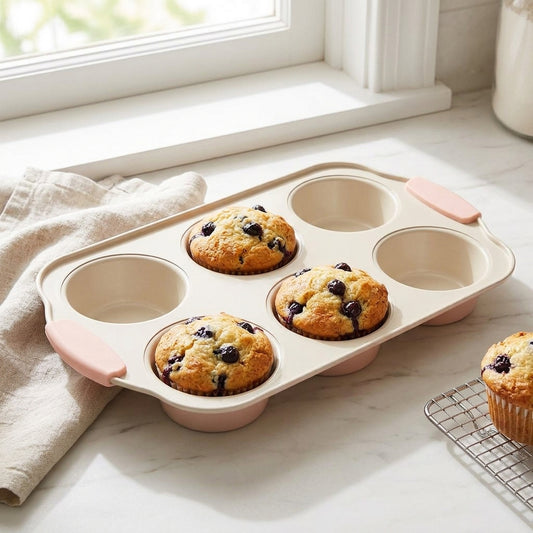 A six-cup muffin tin with pale pink handles and a beige interior, shown on a bright kitchen counter with four baked blueberry muffins. A folded beige cloth and a cooling rack with a muffin are visible near the window.