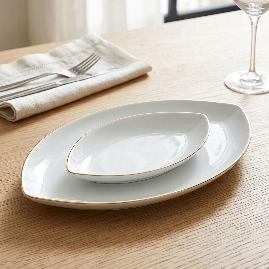 Three nested oval ceramic plates in white with a thin gold rim, arranged on a light wood table next to a folded cream napkin with silverware and a wine glass.