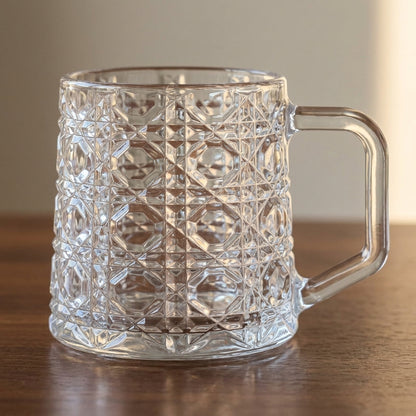 A clear glass mug with a textured diamond-cut pattern and a sturdy handle, resting on a wooden surface against a neutral background.