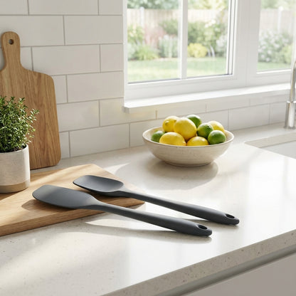 Black spatulas on a kitchen counter with a bowl of fruit and cutting board in the background.