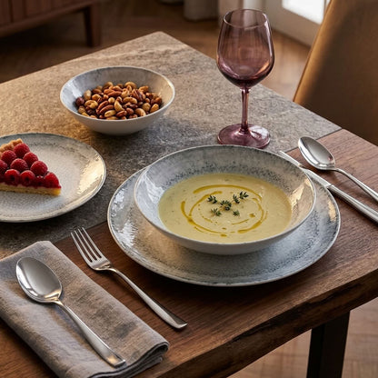 A styled dining table setting featuring a ceramic soup bowl with a speckled blue-gray rim, filled with creamy yellow soup and garnished with olive oil and herbs. The bowl sits on a matching round charger plate.