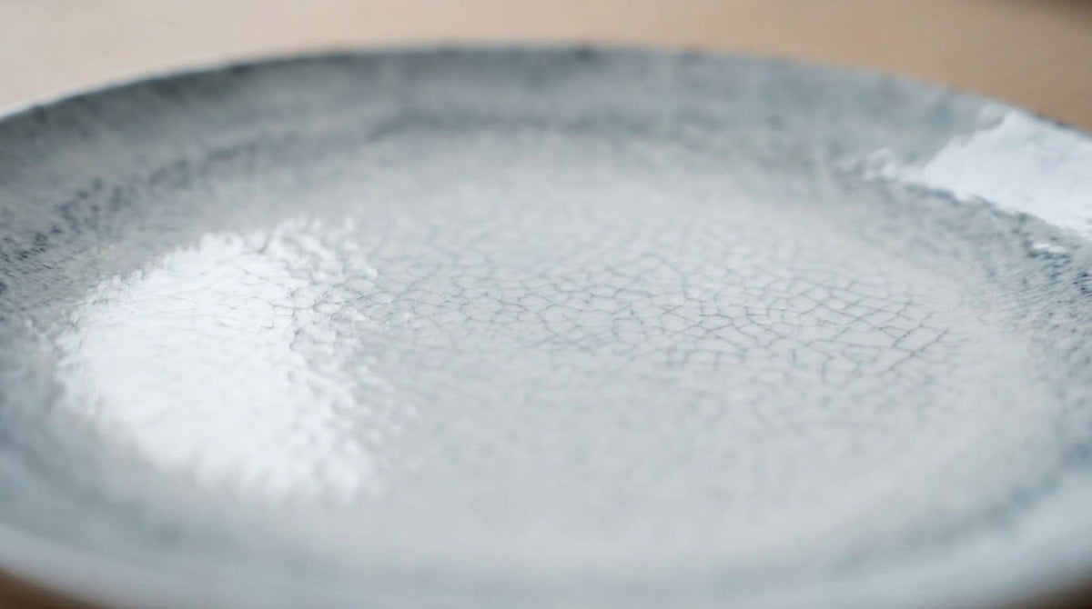 A close-up of a ceramic plate with a textured, crackled glaze in light gray and white, featuring a subtle blue-gray rim.