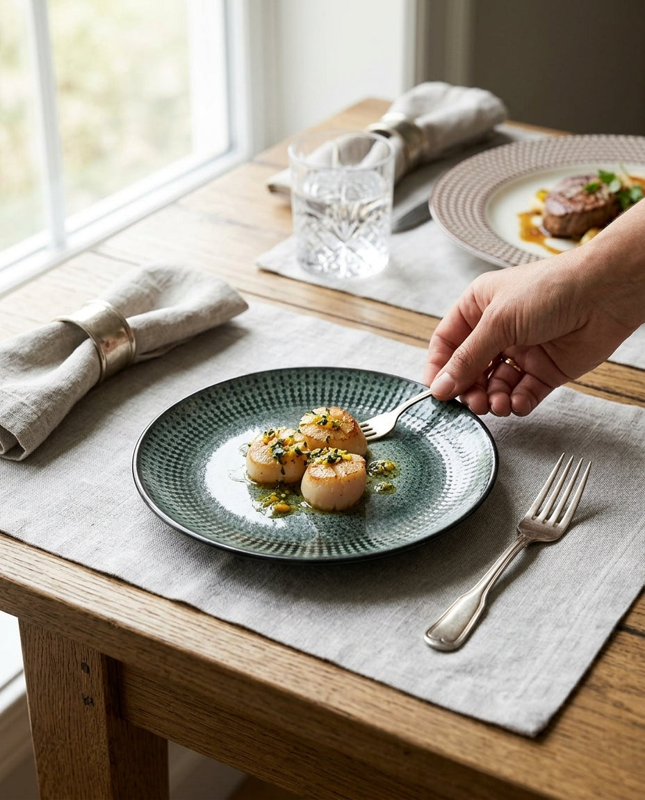 A hand uses a fork to eat from a round textured teal-green plate holding three scallops, set on a light gray placemat on a wooden dining table.
